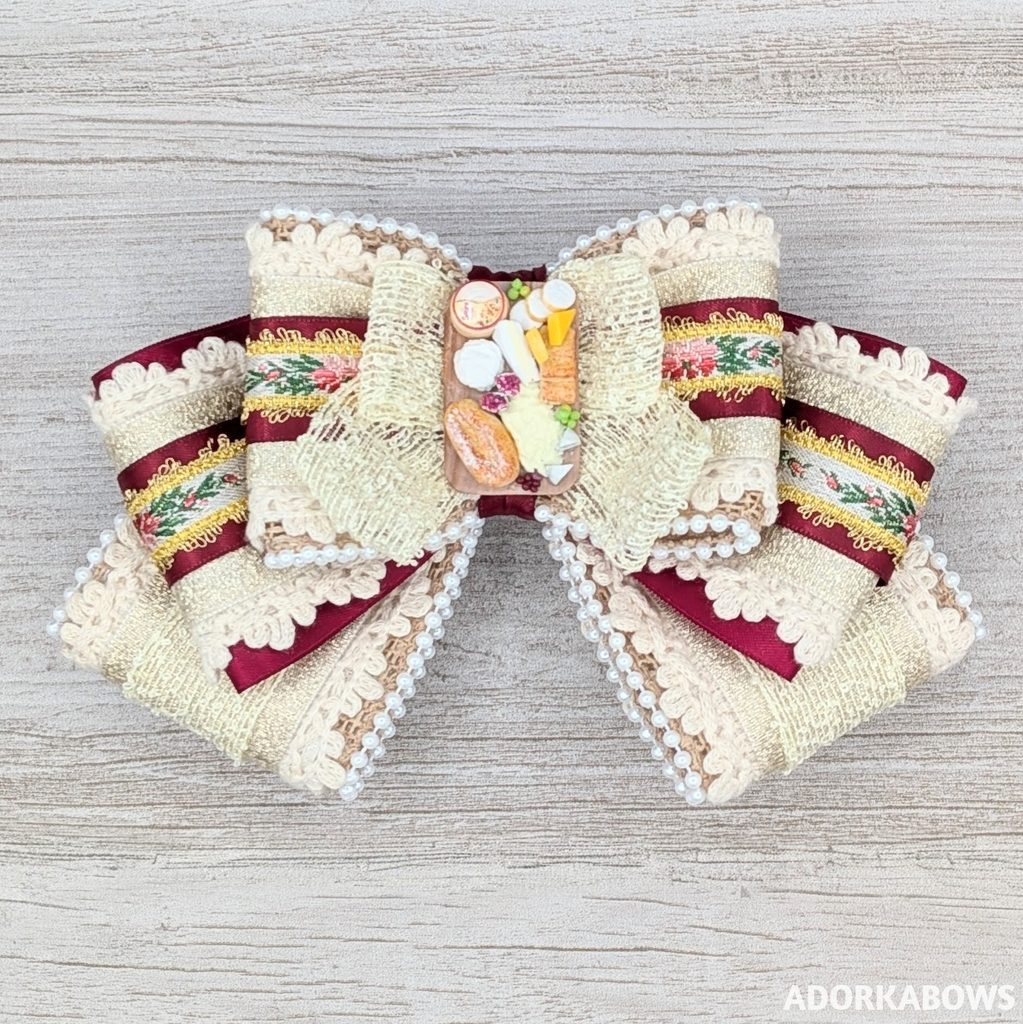 A charcuterie themed hair bow featuring a tiny clay board by BigMadeSmall with accent ribbons in red, floral embroidery, lace and pearls