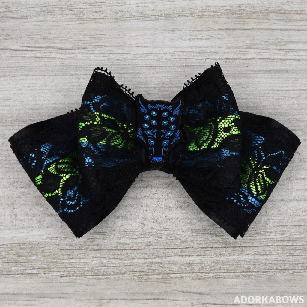 A mostly black hair bow with lace laying over blue and neon green layers; the center is a wolf head with a dozen eyes which glow in the dark. Pin by Aimee Cozza and used with permission.