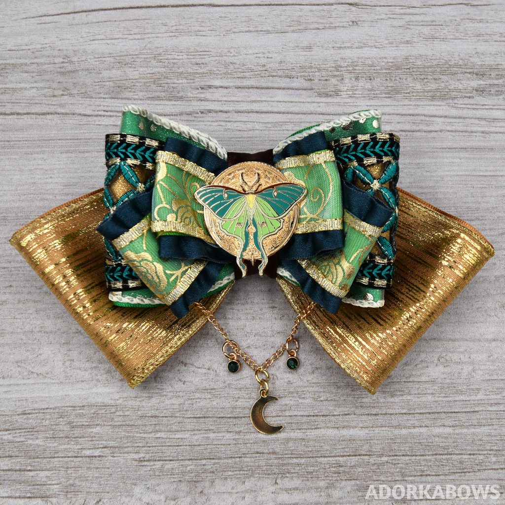 A gold and teal hair bow with green accents, featuring floral and lattice patterns. The center is a round pin of gold with a stunning lunar moth on it in similar colors. Below the bow hangs a chain with green gems and a crescent moon. Pin by Rachel Quinlan and used with permission
