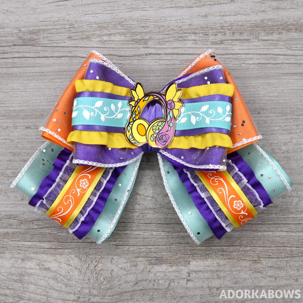 A sparkly, ruffled hair bow in purple, orange, yellow, and minty green with an enamel pin of floral headphones at the center. Pin by me and AmunetGFX and used with permission
