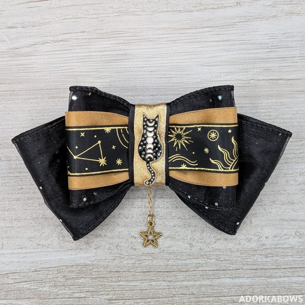 A black and gold hair bow with space metallic patterns featuring a cat pin at the center with the phases of the moon up its back. Pin by AdventureAwaitsME and used with permission