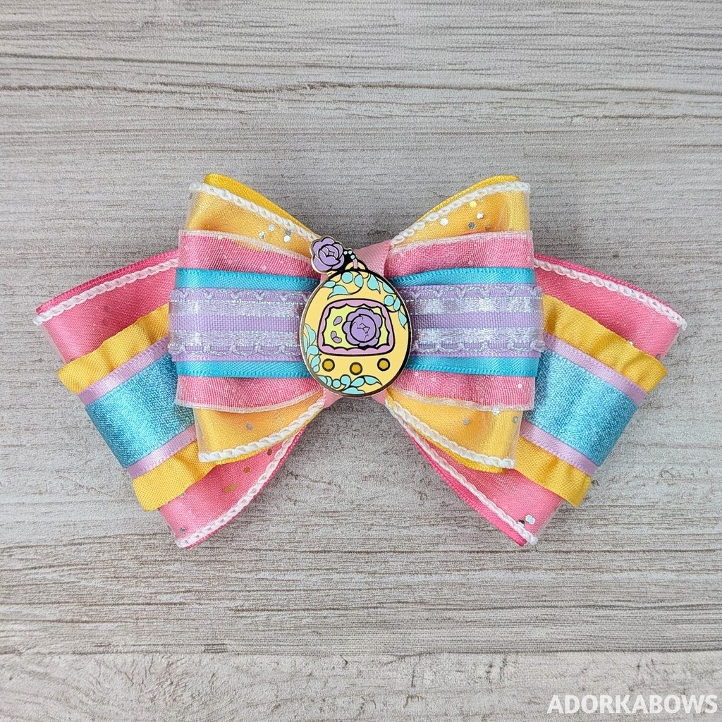 A sparkly hair bow with ruffles and glitter in colors of yellow, pink, aqua, and light purple with a center pin of a floral interpretation of a digital pocket pet. Pin by me and AmunetGFX and used with permission.