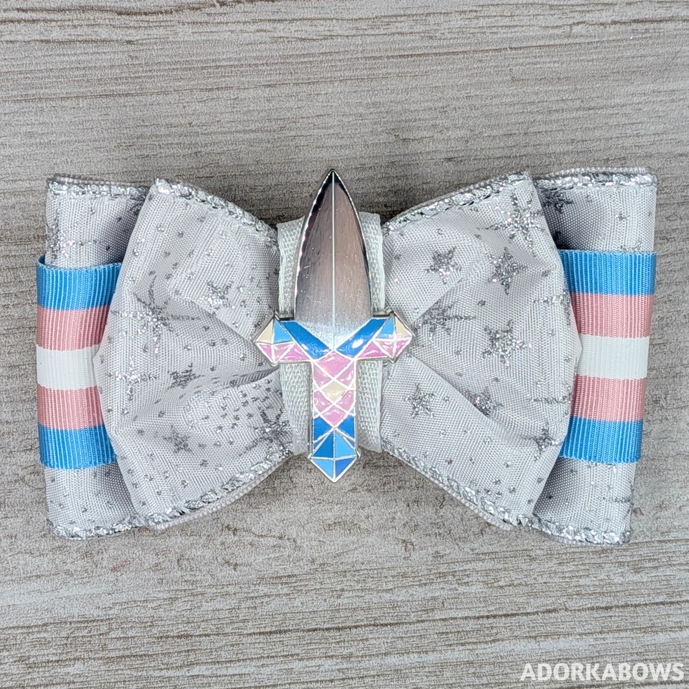 A blue, white, and pink hair bow featuring a sword with trans pride colors. The ribbons are ruffle and sheer and there's a loop behind the pin to make the bow into a convertable keychain/bag hanger. Pin by MarachiStudio and used with permission.