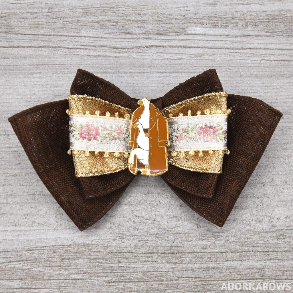 A brown hair bow with gold accents and a center ribbon of floral embroidery; the center pin is three geese in a trenchcoat. The pin is by SugarLich and used with permission.
