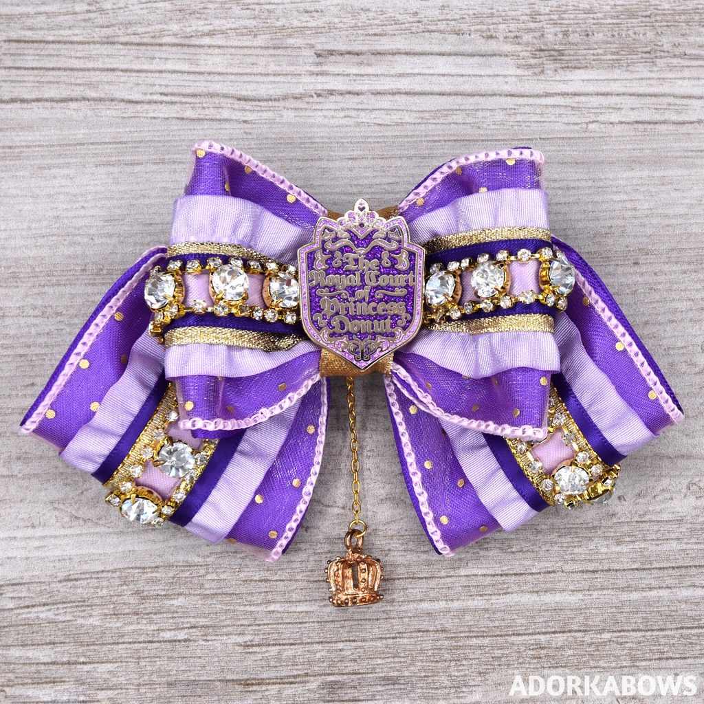 A purple and gold hair bow featuring ruffles and white gems and glitter with a center shield pin that says Royal Court of Princess Donut and a golden charm hanging below the center.