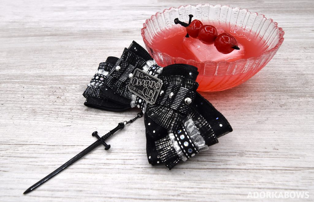 Desperado Club enamel pin from Foam Brain Games made into a one of a kind hair bow with silver and black ribbons, studs, and a dangling black blade. It's resting in front of a glass bowl of red iced liquid with a sword and three cherries on the ice cube. The pin is by Foam Brain Games and used with permission